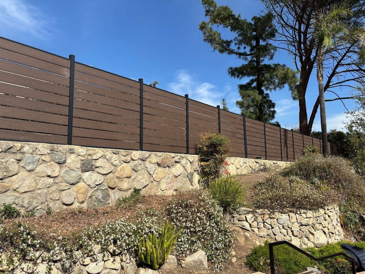 Long brown composite privacy fence on a stone retaining wall above a planted slope