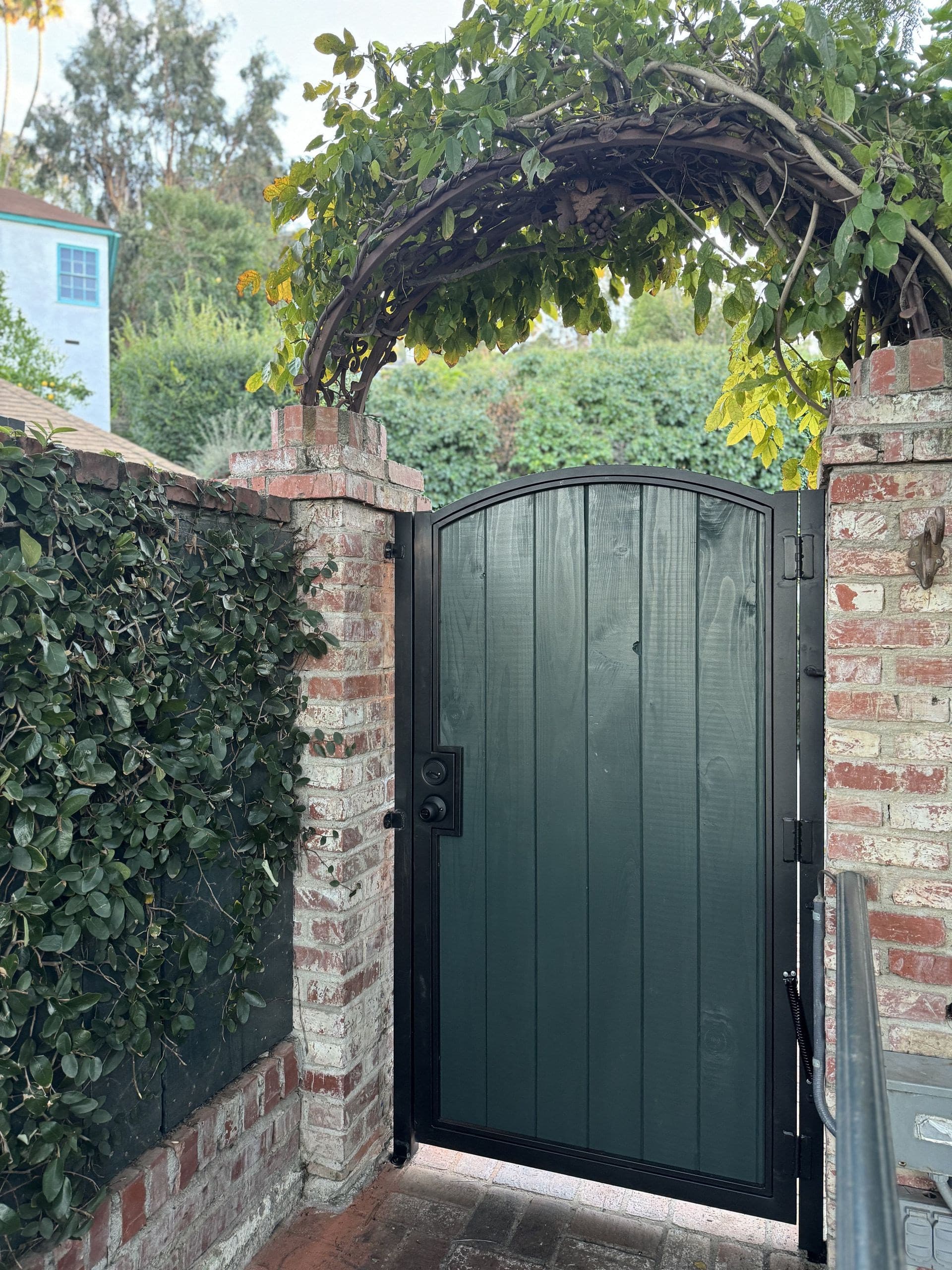Dark green wood gate in a black steel frame with an arched top, between old brick pillars under a vine arch