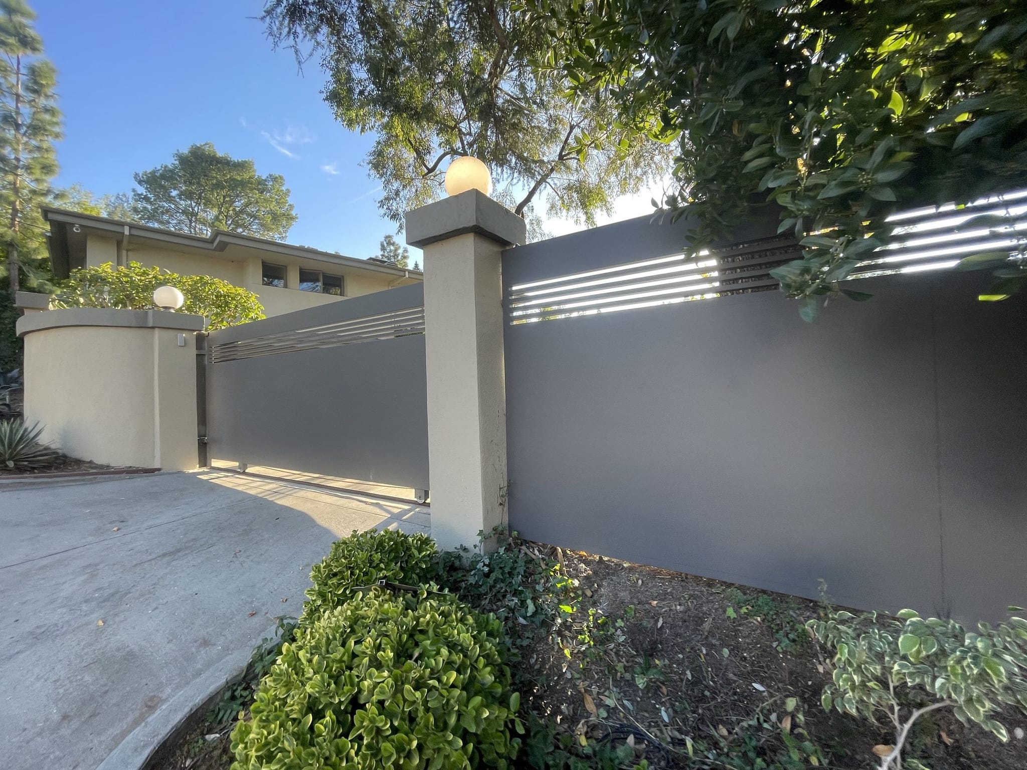 Gray sliding driveway gate and solid fence with slatted tops, between stucco pillars with globe lights