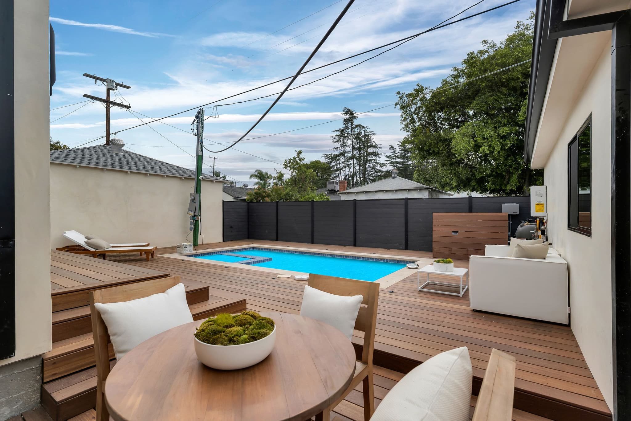 Dining set on a hardwood deck beside a pool, a dark privacy fence along the back of the yard