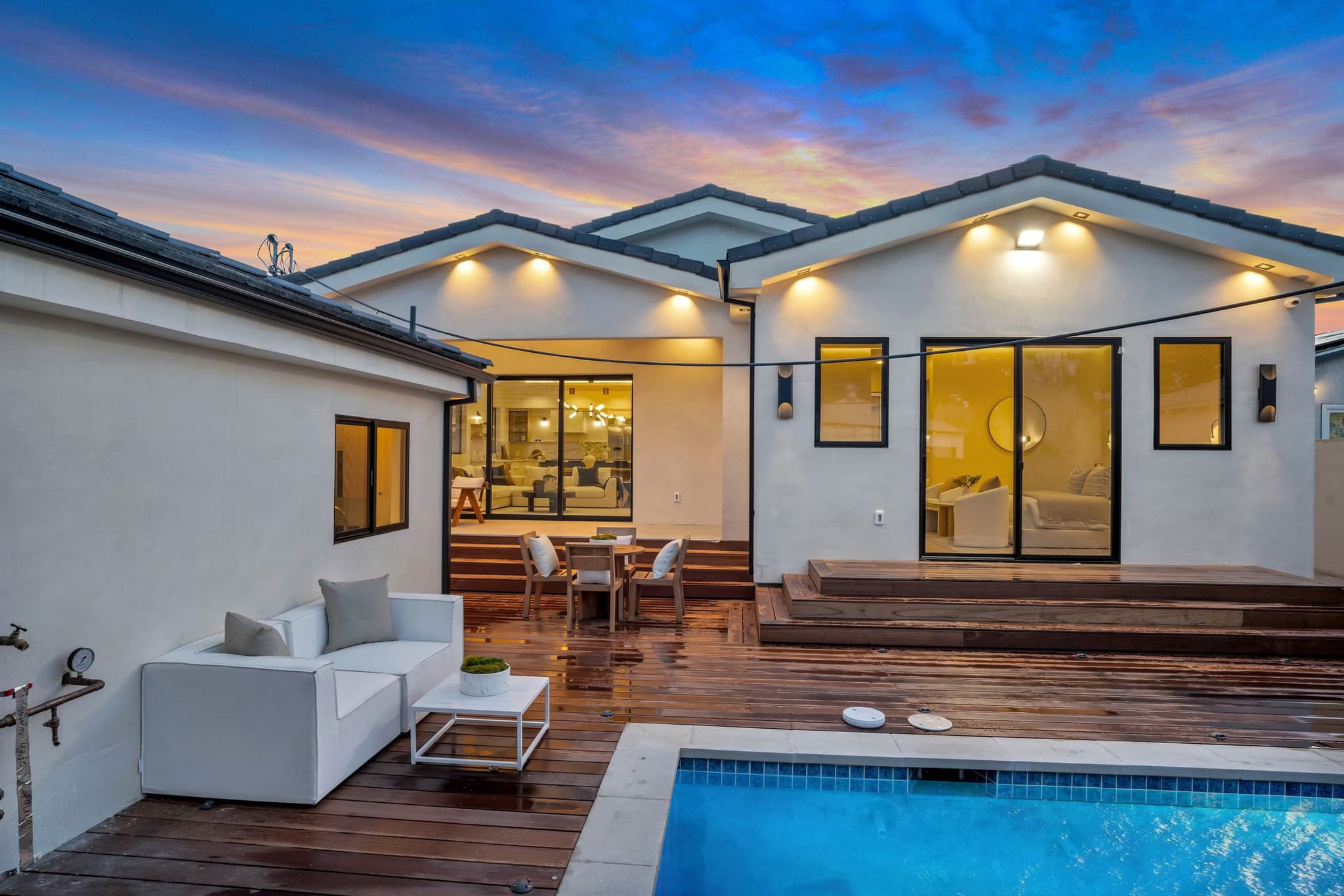 Hardwood deck and pool behind a new home at dusk, lounge seating and the lit kitchen through open glass doors